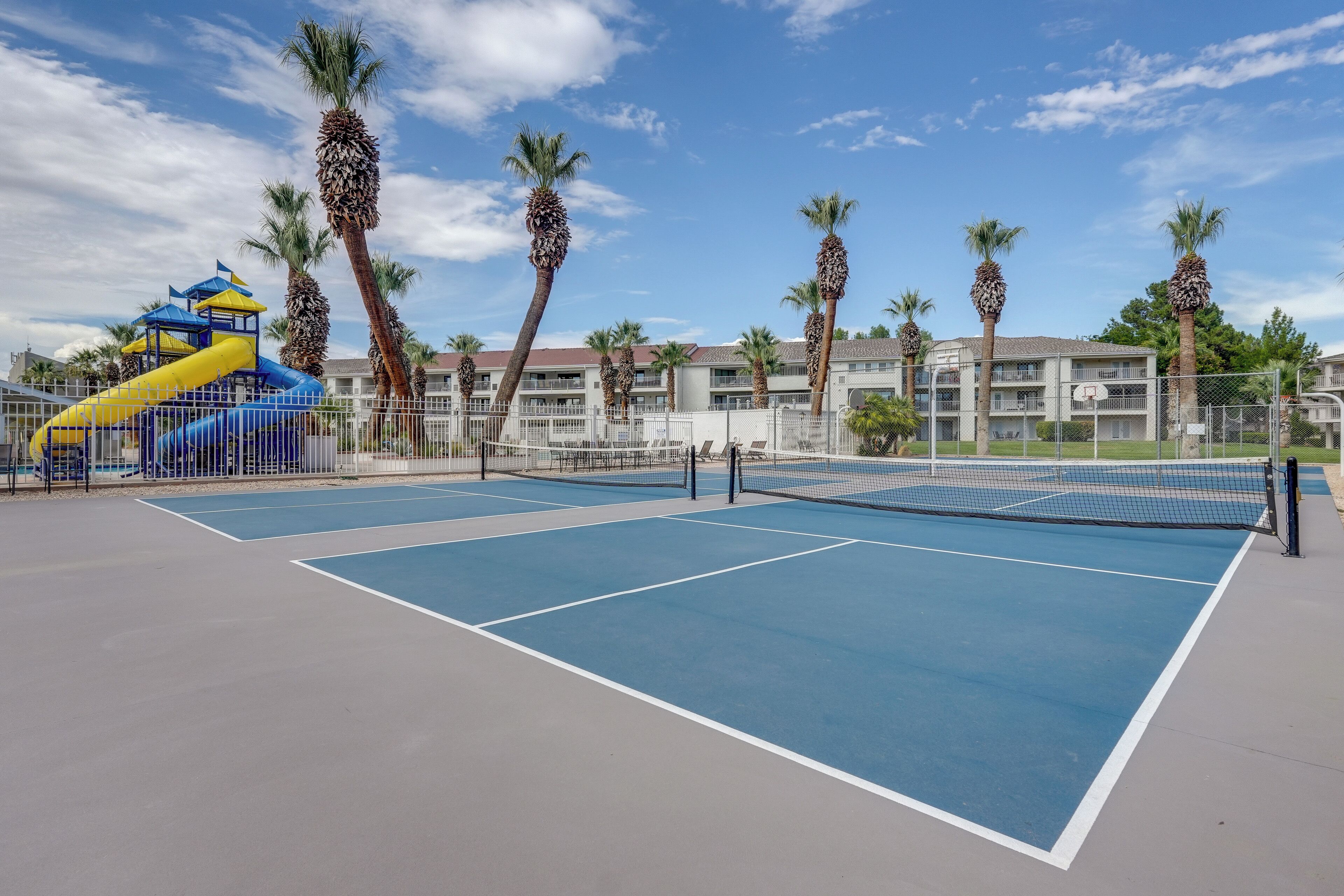 A tennis court with a net, surrounded by palm trees and a playground with slides.