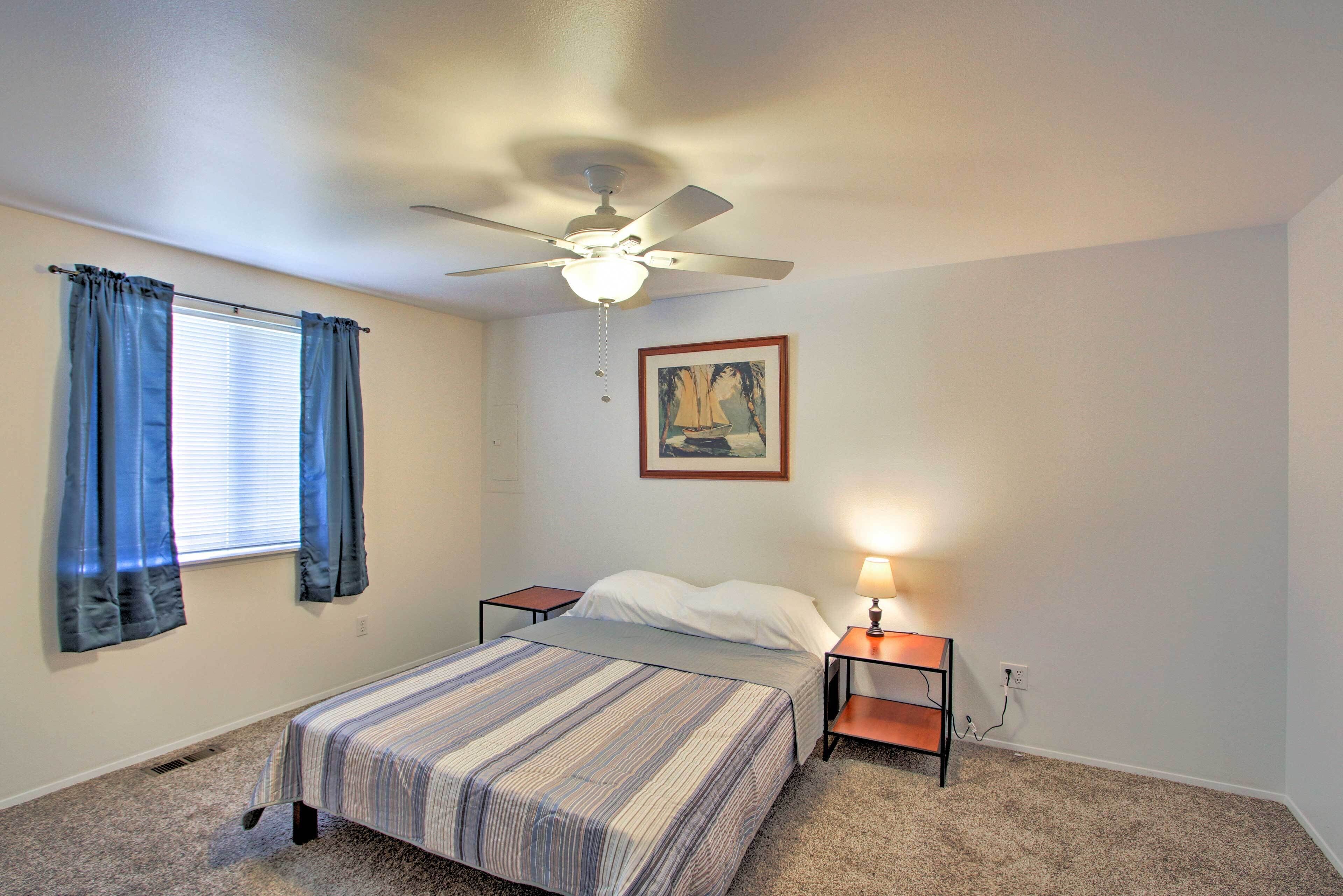 A bedroom with a single bed, a ceiling fan, a nightstand with a lamp, and a window with curtains.
