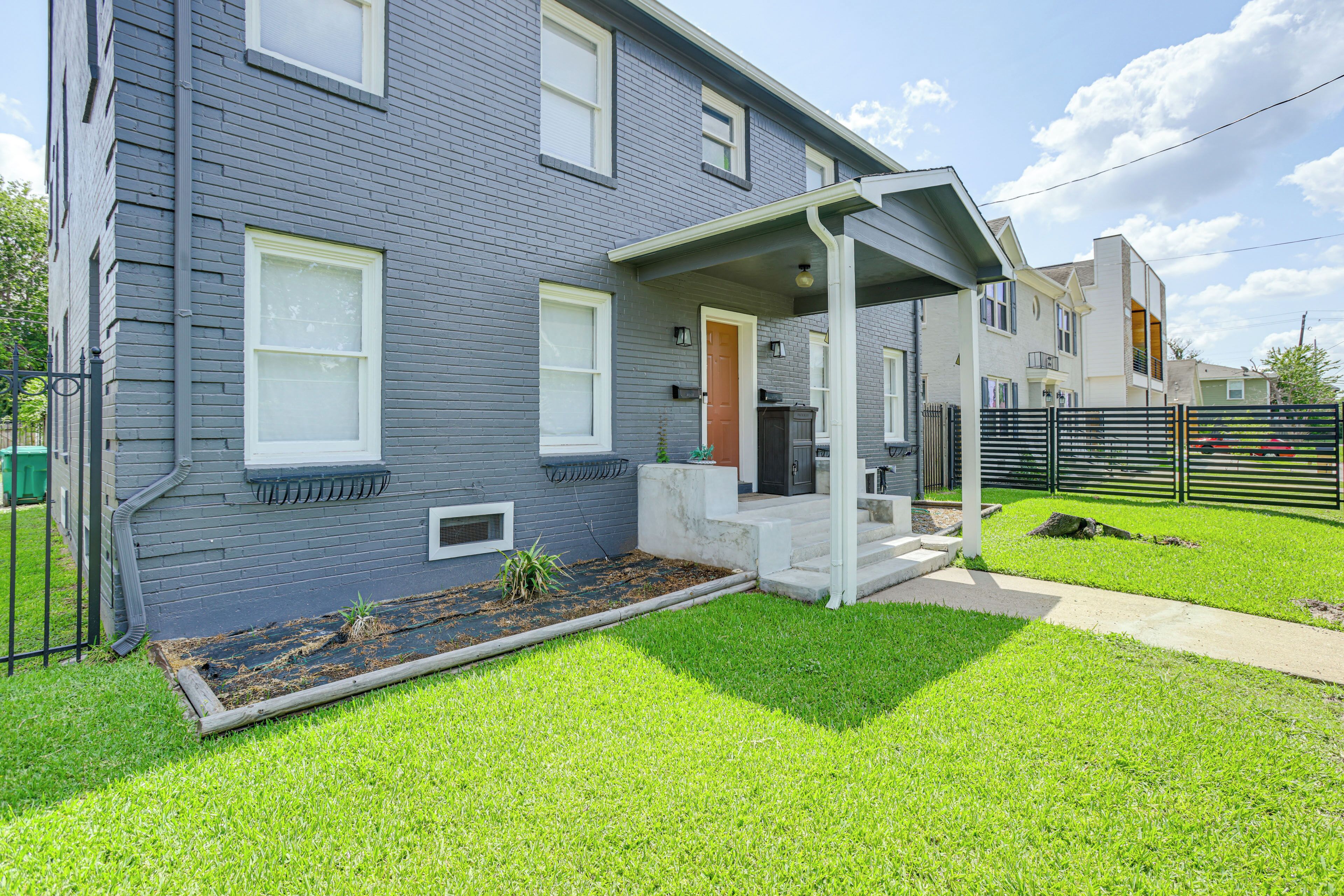 A two-story residential building with a covered porch, a small front yard with a garden bed, and a visible front door.