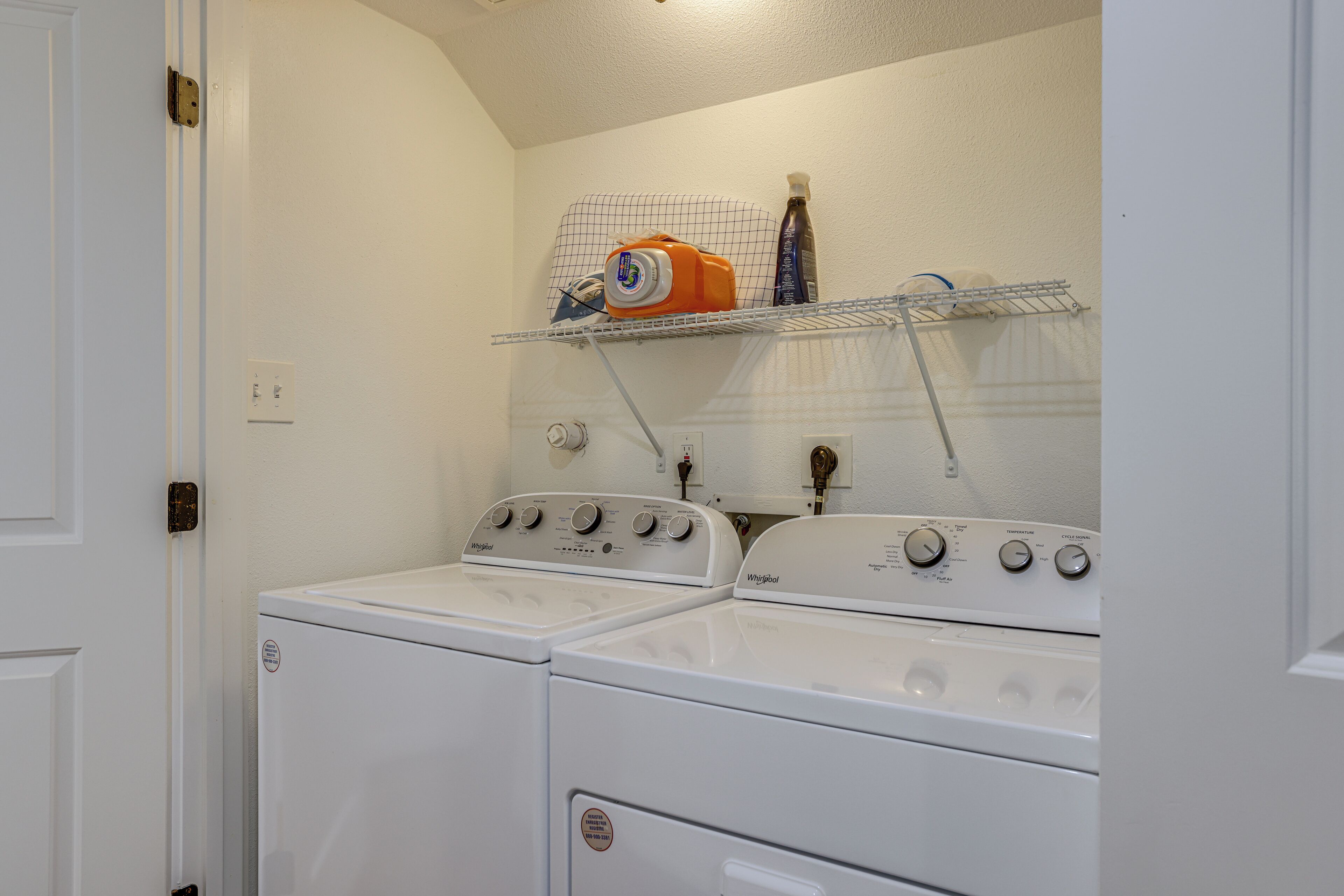 A laundry room with two stacked washers and dryers, a shelf with cleaning supplies, and a wall-mounted heater.