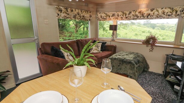 A dining area with a table set for two, a sofa, and a window with a view of greenery. - Gwybedog (Llanwrda)