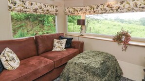 A room with a brown sofa, floral-patterned curtains, a lamp, and a view of greenery outside. - Gwybedog (Llanwrda)