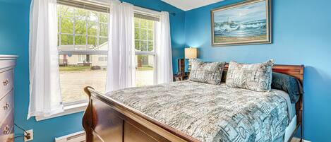 A bedroom with a bed, a dresser, a window with curtains, a painting on the wall, and a lamp on a nightstand.