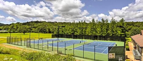 A tennis court with a net, surrounded by a fence, and a building with a chimney in the background.