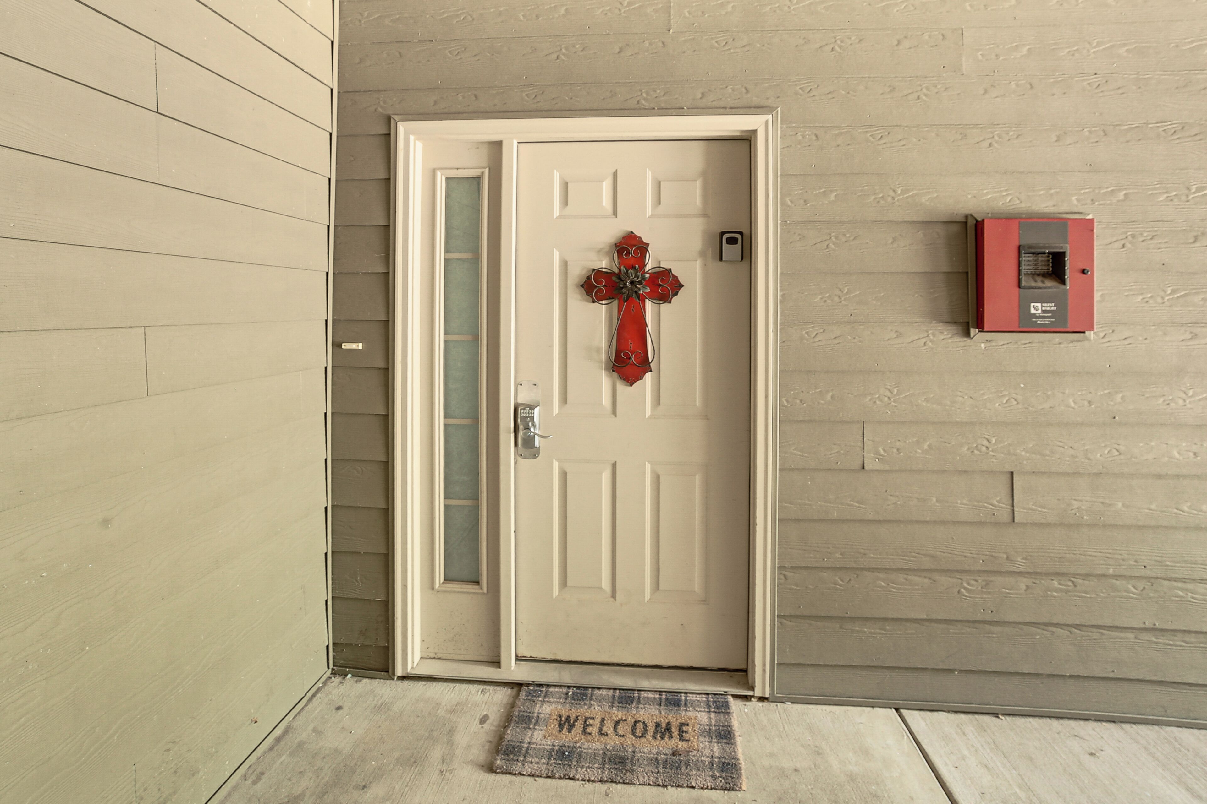 A beige wooden door with a red cross decoration, a doormat with "WELCOME" text, and a red electrical box on the wall.
