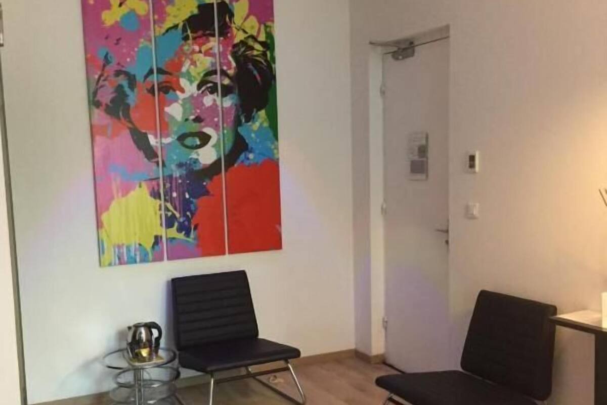 A modern hotel room with a large bed, two black chairs, a small table with a kettle, and a colorful abstract painting on the wall.