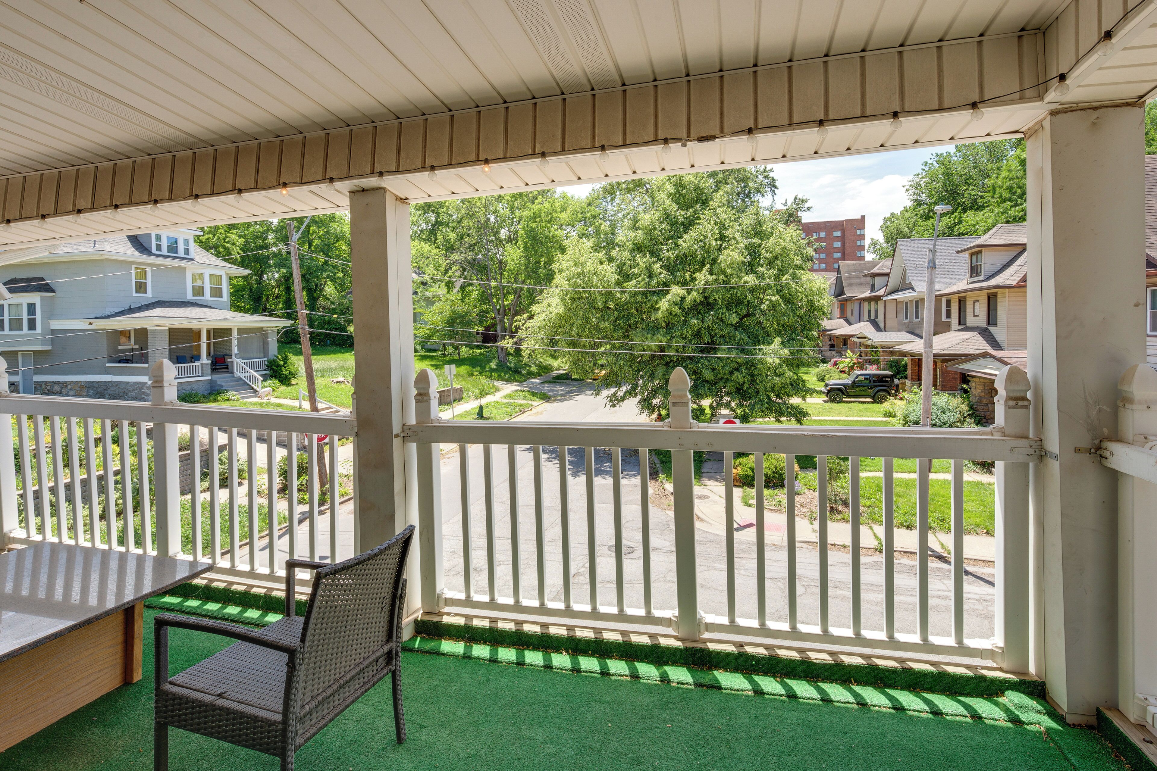 A covered porch with a green artificial turf floor, white railing, and a view of a residential street with houses and a tree.