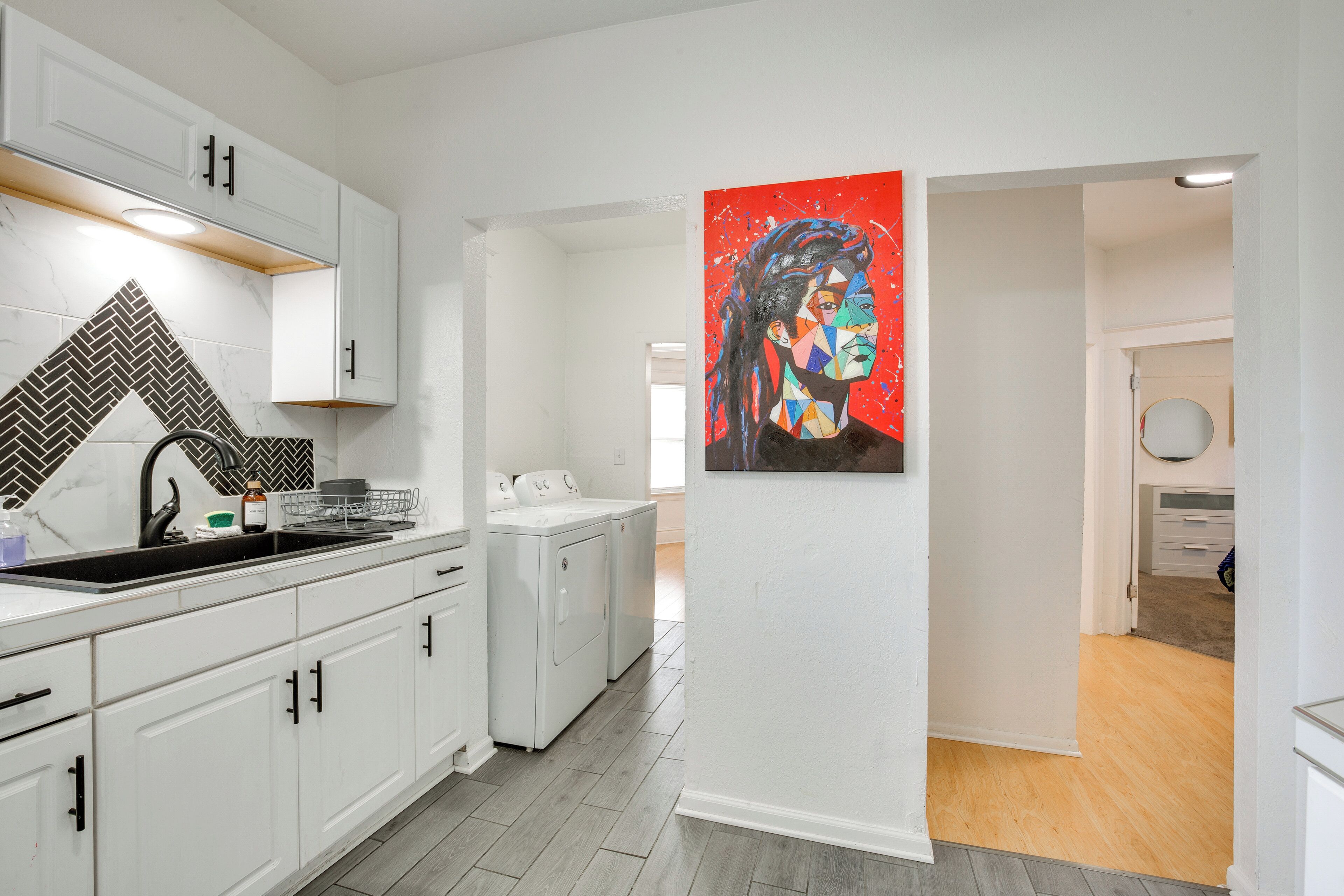 A modern kitchen with white cabinetry, a herringbone tile backsplash, and a large painting on the wall.