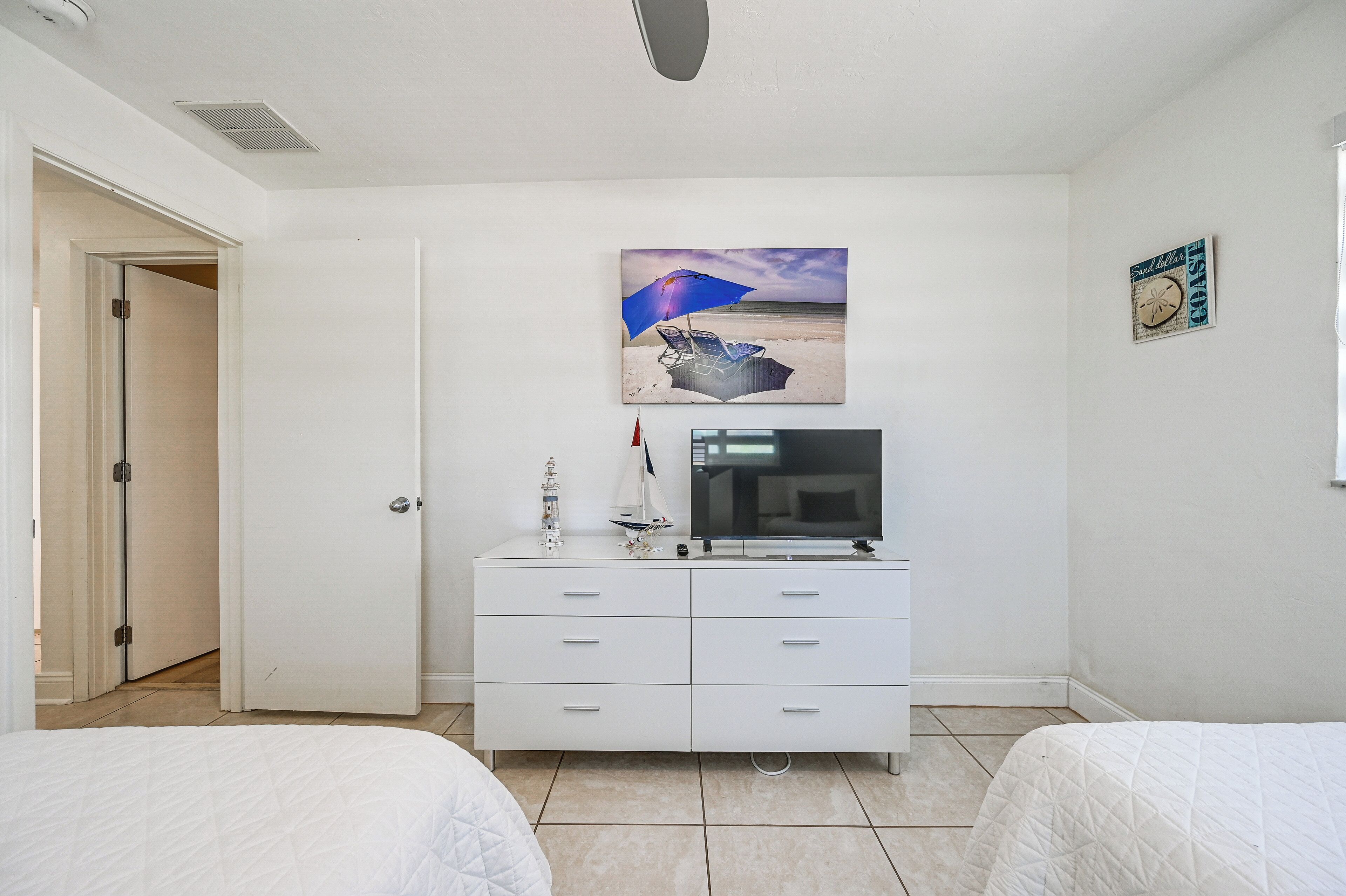 A bedroom with two beds, a white dresser, a television, and a beach-themed painting on the wall.