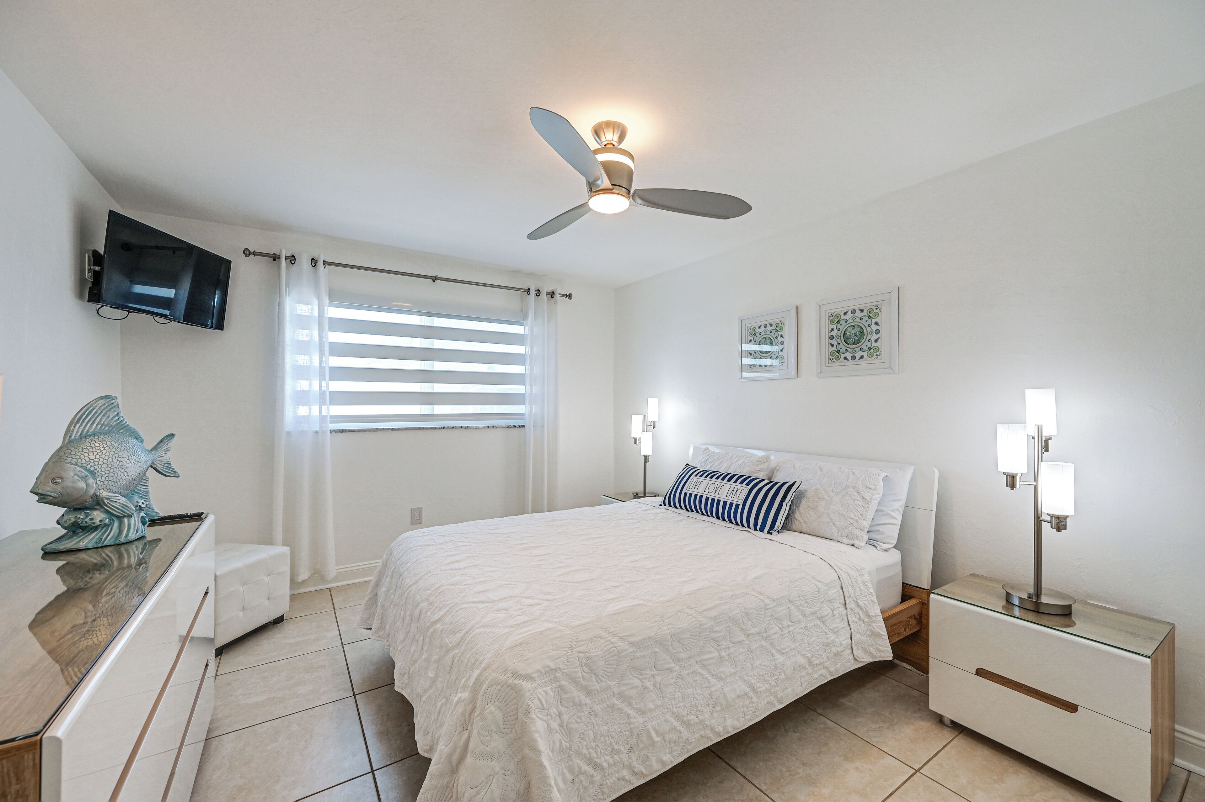 A bedroom with a bed, a ceiling fan, a TV mounted on the wall, a nightstand with a lamp, and a window with blinds.
