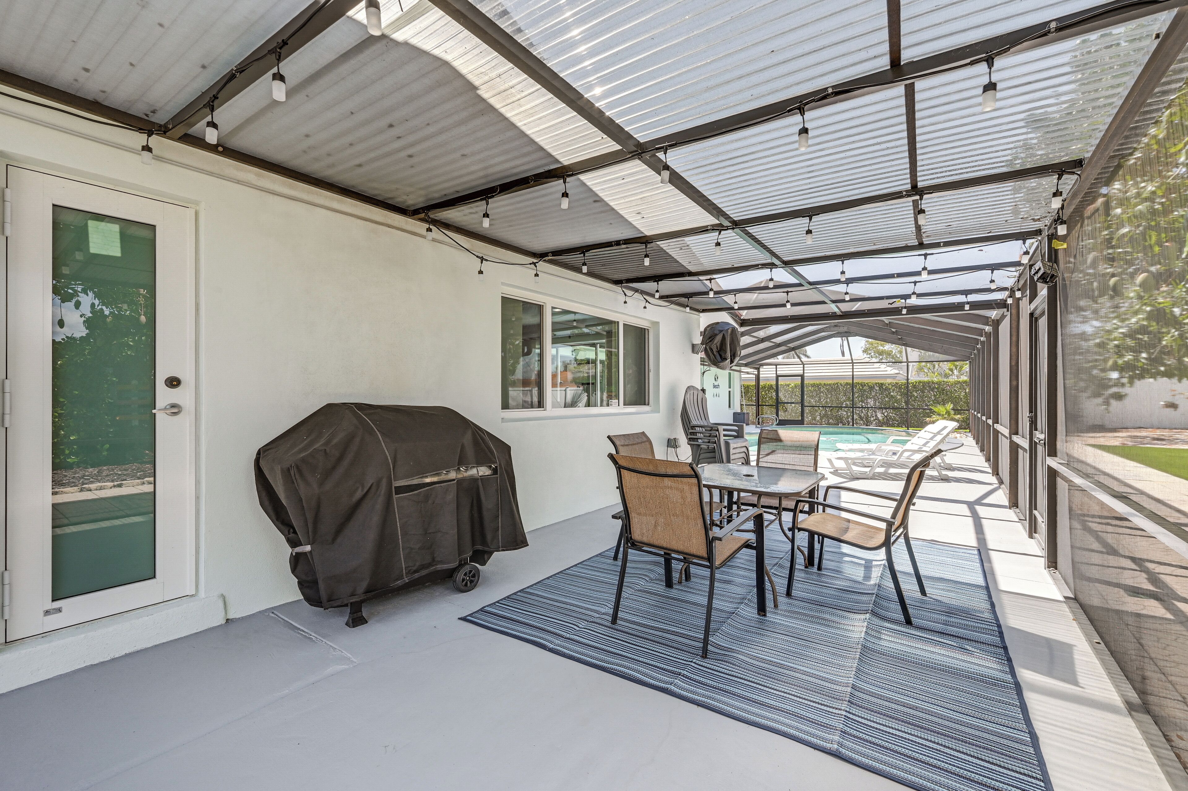 A covered patio with a table and chairs, a grill, and a view of a swimming pool.