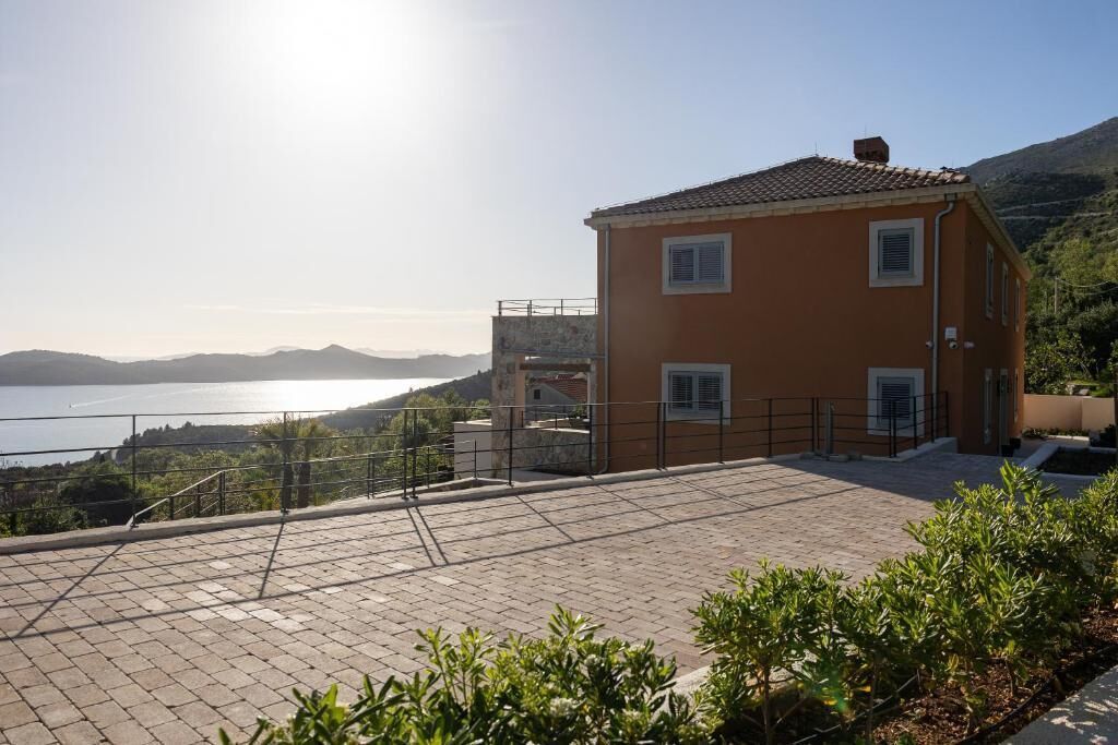 A two-story building with a tiled roof, a balcony, and a view of a body of water and hills in the background.