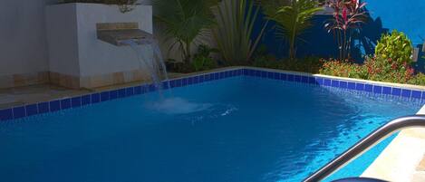 Una piscina con borde de azulejos azules, una fuente de agua y plantas tropicales alrededor.