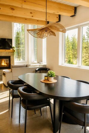 Een moderne eethoek met een grote tafel, zwarte stoelen, een geweven hanglamp, een open haard en grote ramen met uitzicht op bomen.