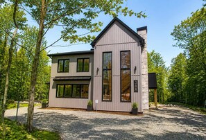 A two-story house with a stone chimney, large windows, and a gravel driveway. - Spa & Lake | Orphee (Chertsey)