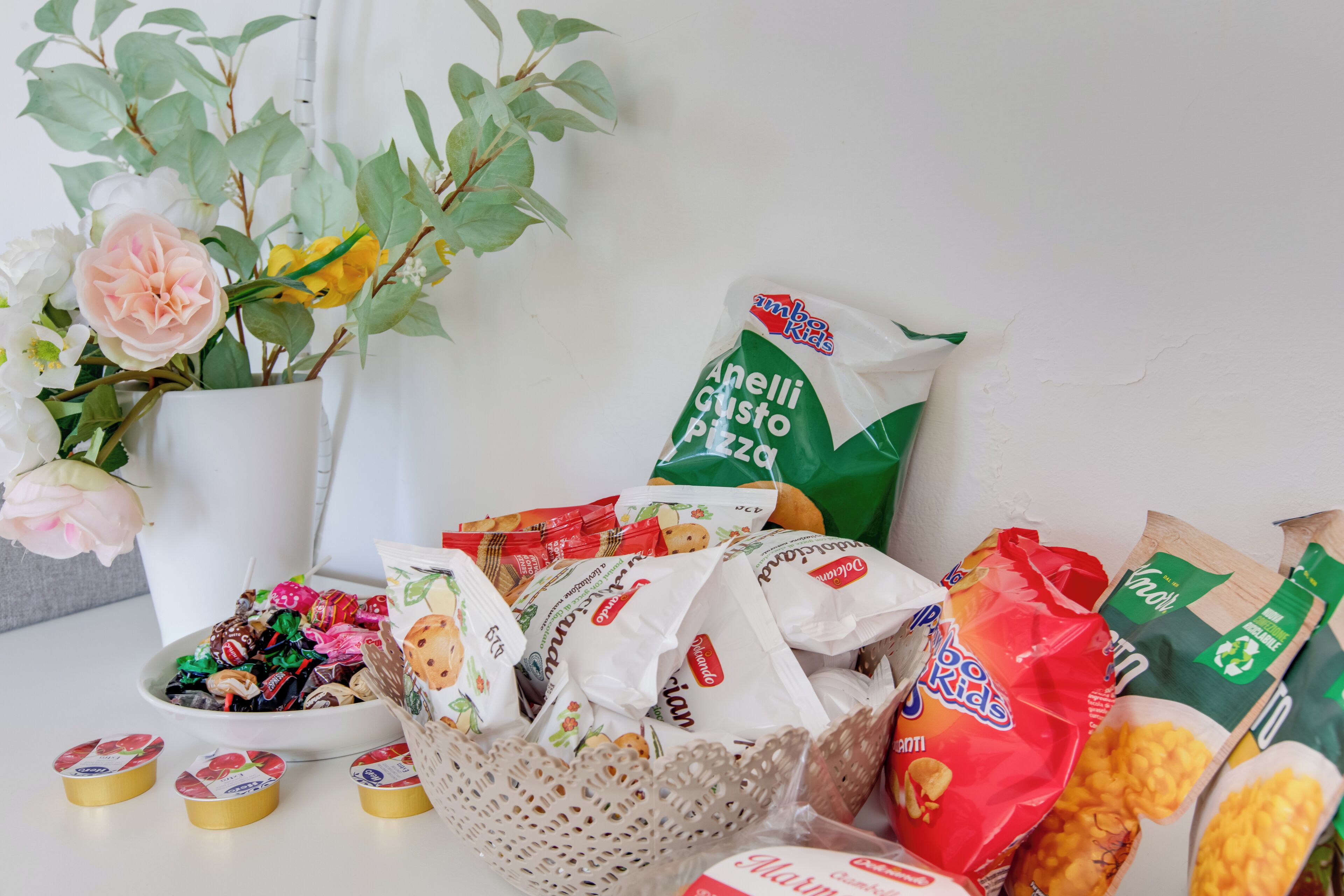 A table with various food items, including a basket of snacks, a vase with flowers, and several packaged goods.