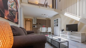 A modern living room with a brown sofa, a glass coffee table, and a TV on a white cabinet. A kitchen with white cabinets and a dining area with white chairs is visible in the background. - Loft Cioccolato by Wonderful Italy (Turin)