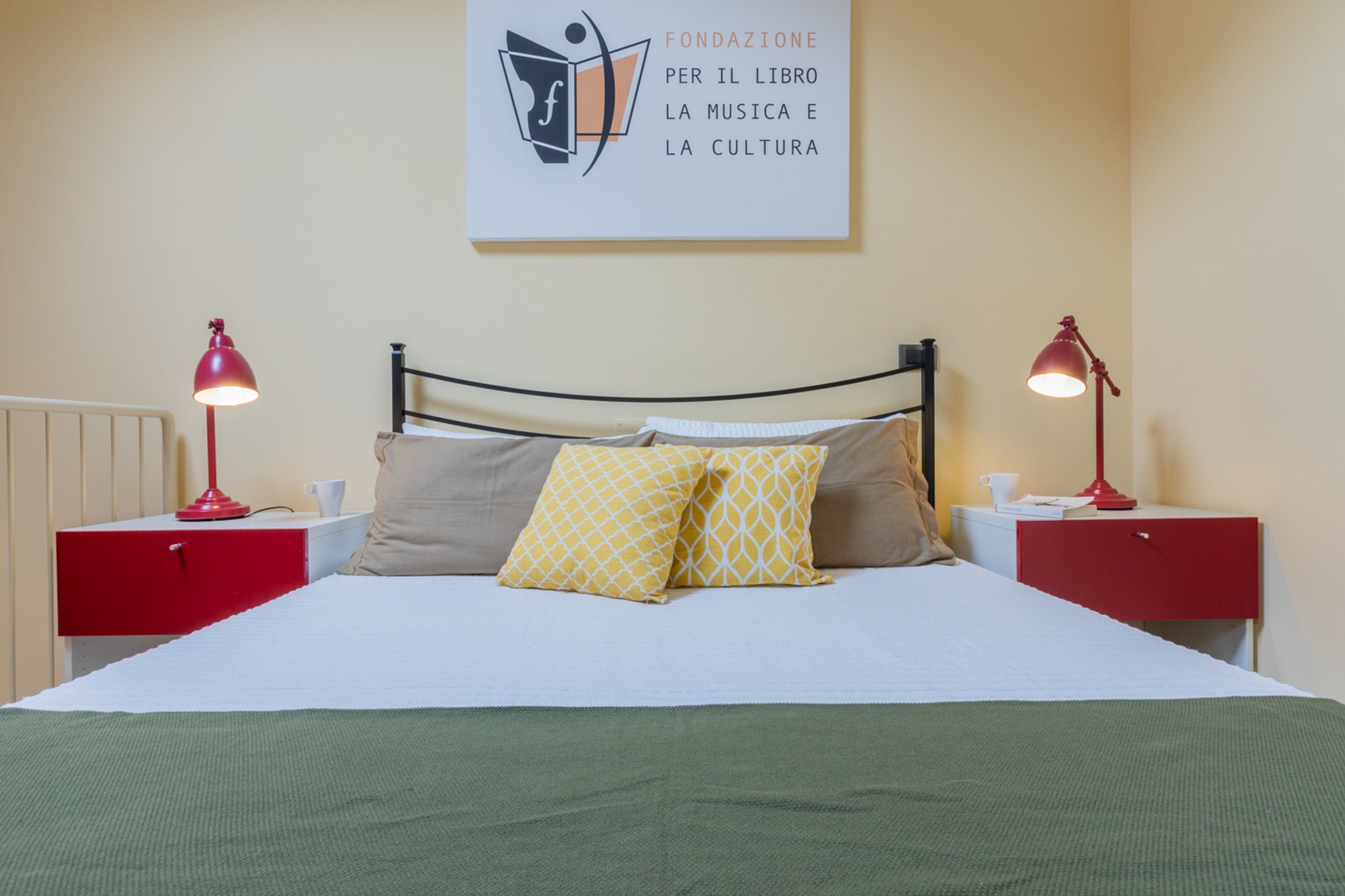 A neatly made bed with two pillows, a red bedside table with a lamp, and a wall with a framed sign.