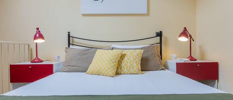A neatly made bed with two pillows, a red bedside table with a lamp, and a wall with a framed sign.