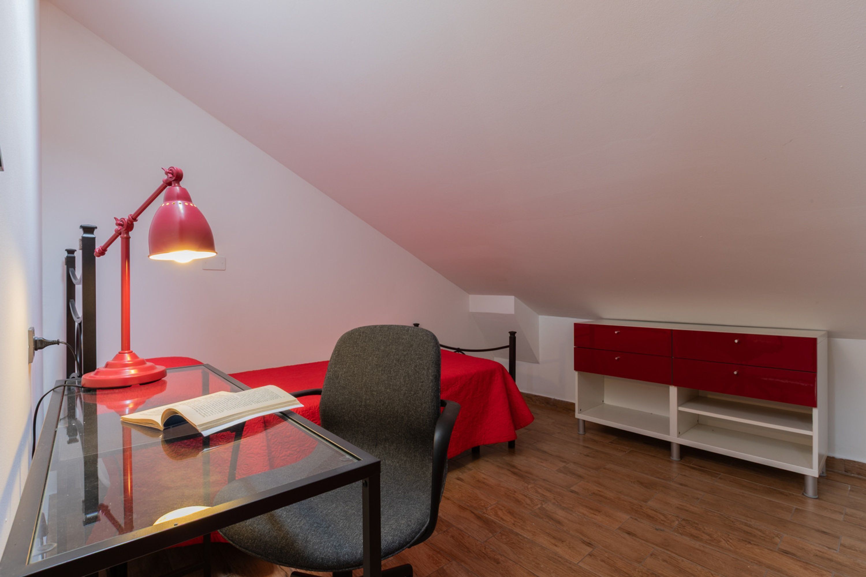 Une pièce comprenant un bureau en verre, une lampe de bureau rouge, une chaise, un lit recouvert d’une couverture rouge et une commode blanche.