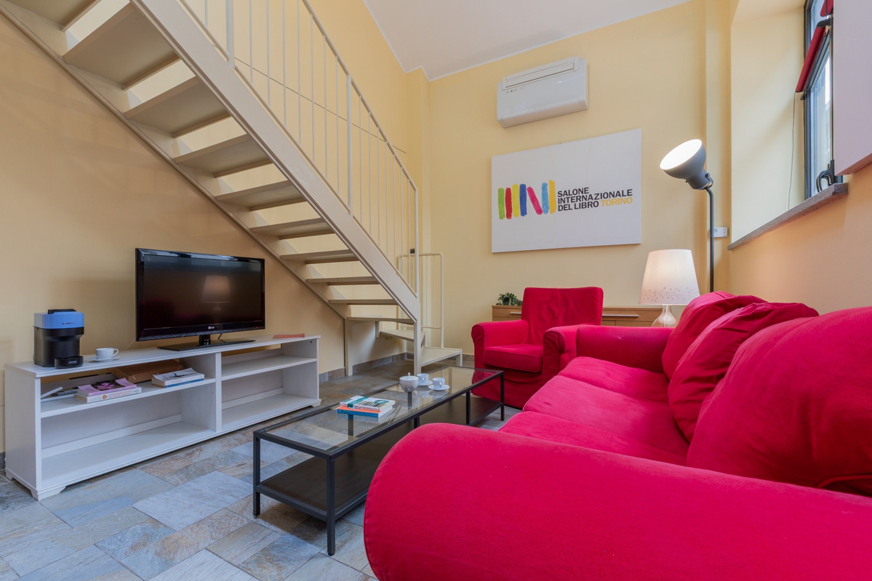 A living room with a staircase, a flat-screen TV, a red sofa, a coffee table, and a bookshelf.