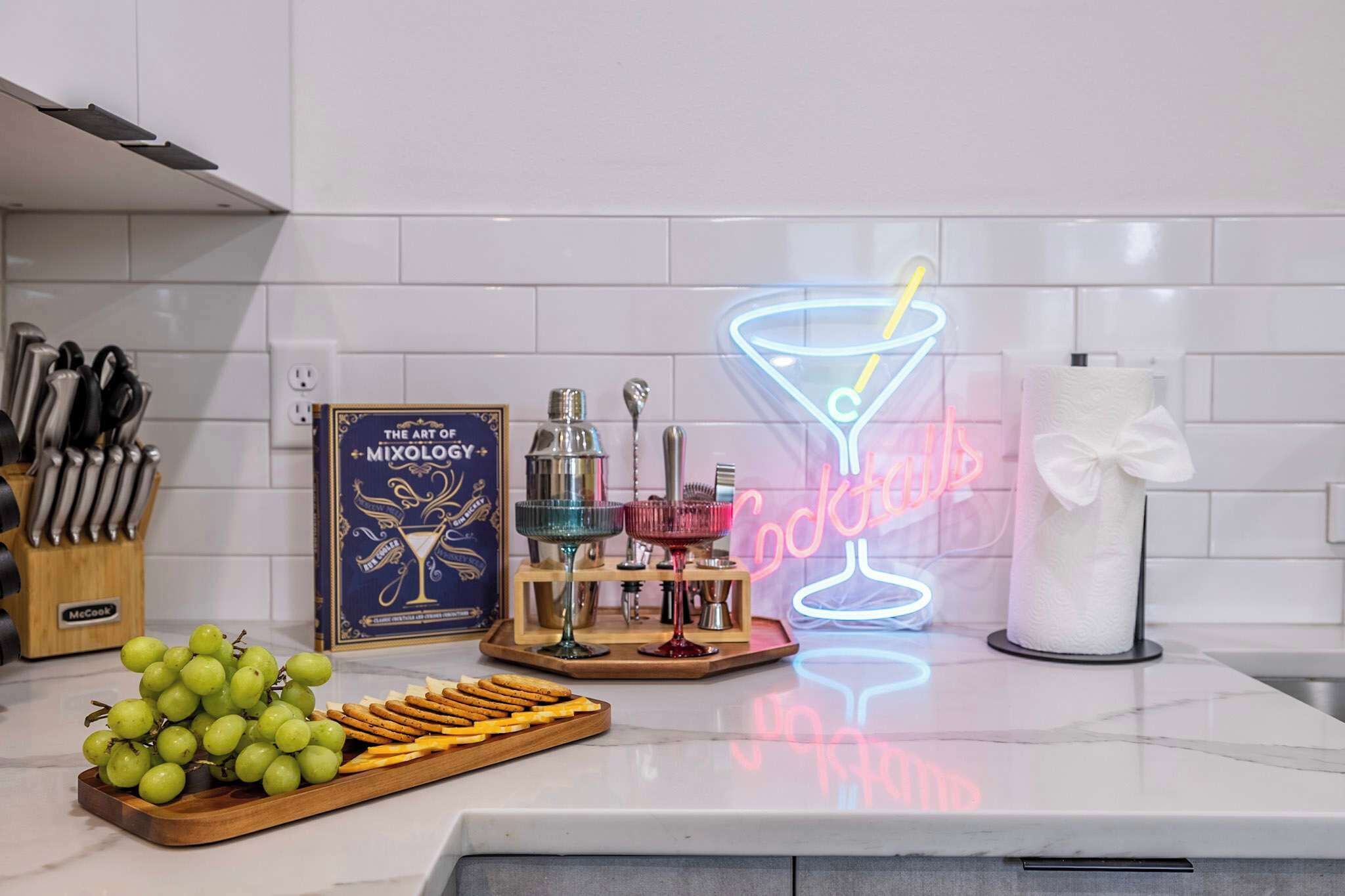 A kitchen counter with a neon cocktail sign, a framed book, a bottle of alcohol, a tray of grapes and crackers, and a paper towel holder.