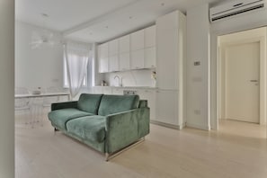 A modern living room with a green sofa, a dining area with a white table and chairs, and a kitchen with white cabinets and a white countertop.