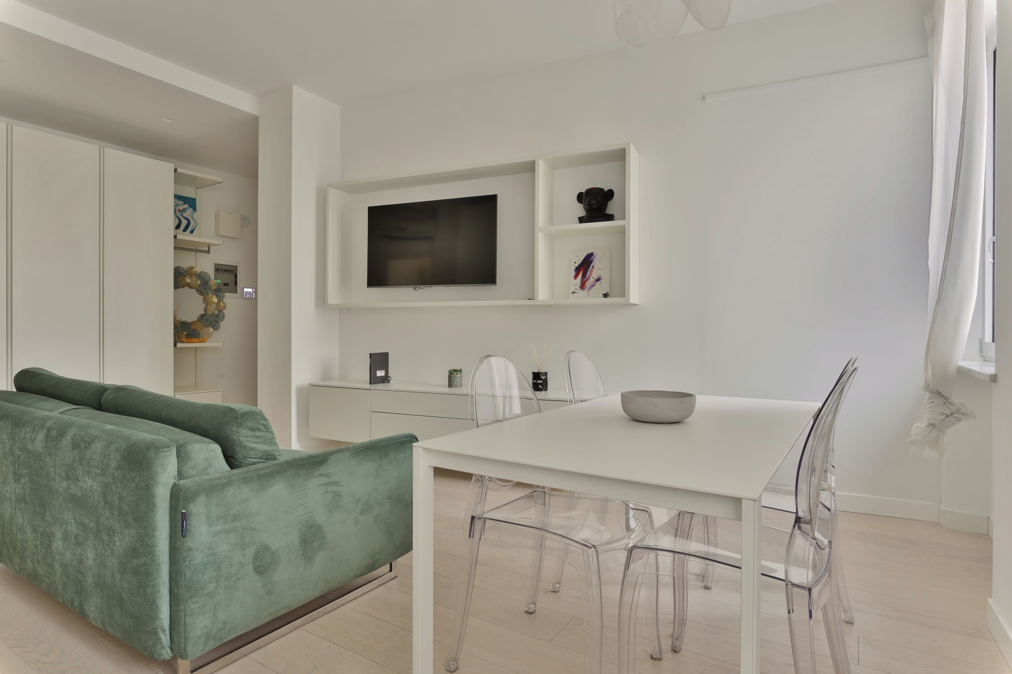 Ein modernes Wohnzimmer mit einem grünen Sofa, einem weißen Esstisch mit transparenten Stühlen und einem an der Wand befestigten Flachbildfernseher.