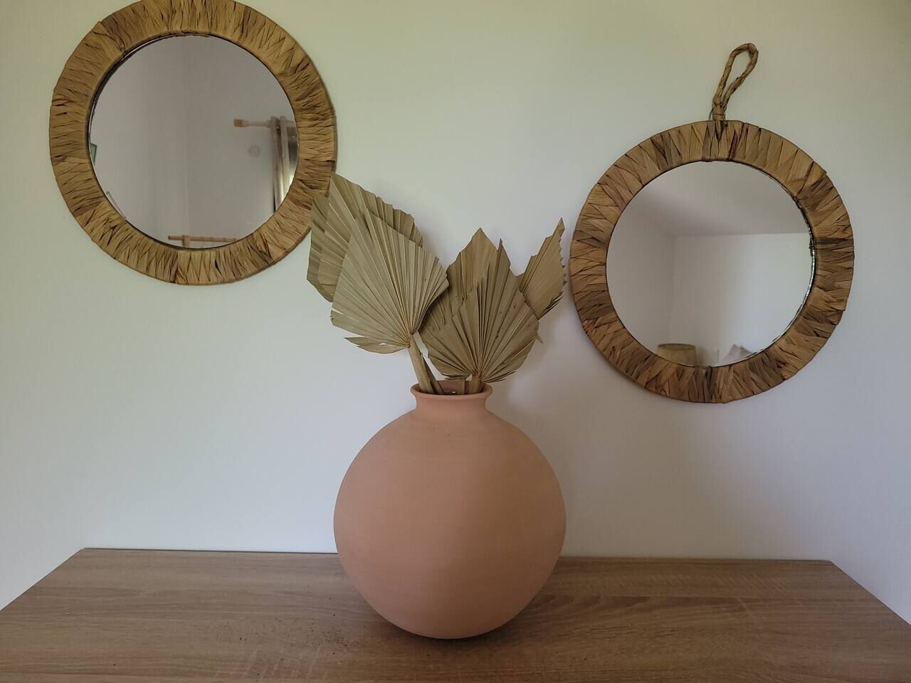 Un jarrón con hojas secas sobre una mesa de madera, frente a una pared blanca con dos espejos redondos.