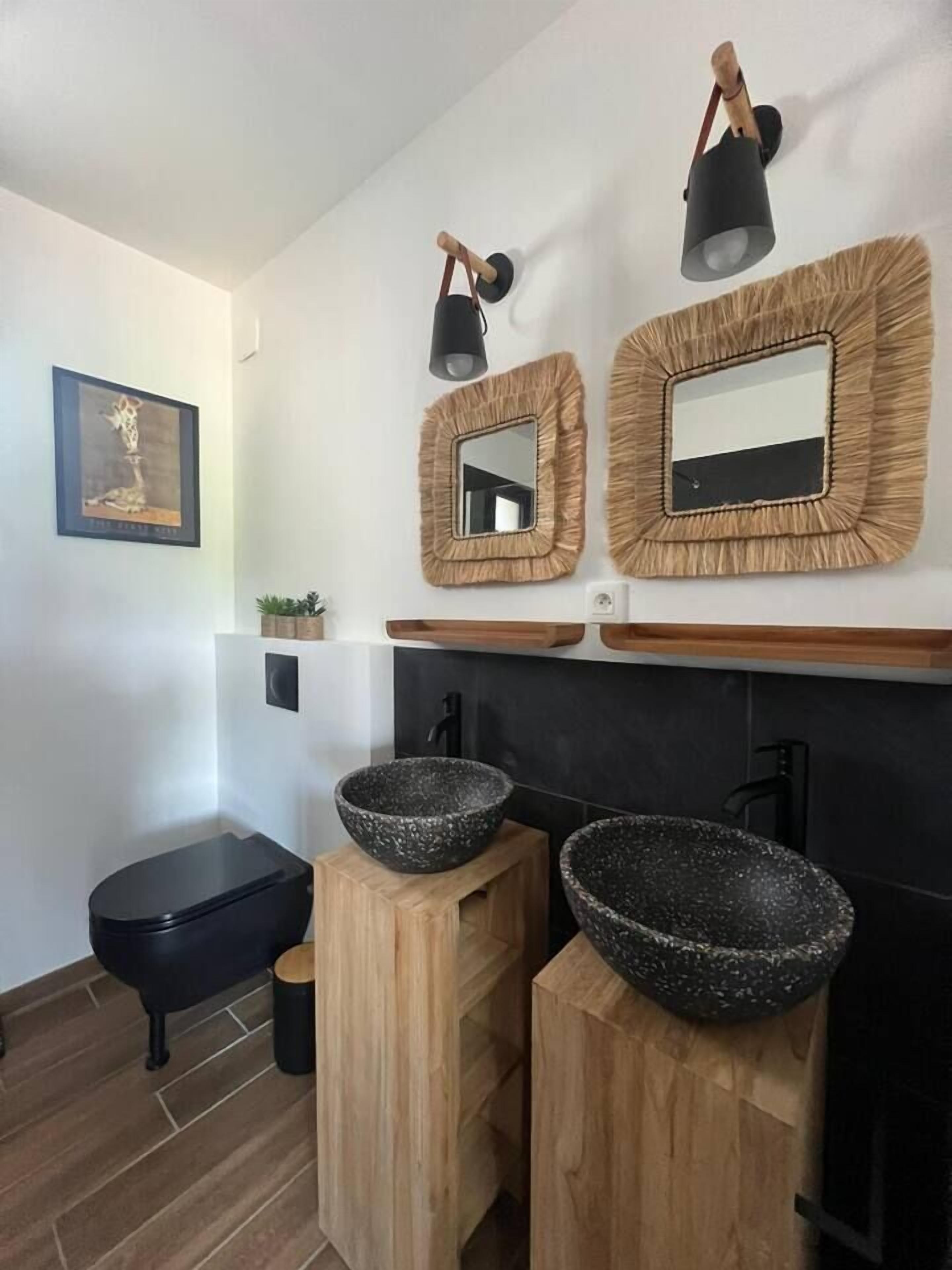 Ein Badezimmer mit einer schwarzen Toilette, zwei hölzernen Hocker mit schwarzen Schüsseln, einem Spiegel mit gewebtem Rahmen und einer wandmontierten Leuchte.