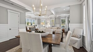A spacious dining area with a wooden table and white upholstered chairs, a chandelier, and a view of a kitchen and dining area through large windows. - Surf Sanctuary + Golf Cart, Pet Friendly & Bikes (Santa Rosa Beach)
