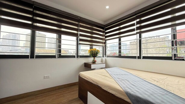Un dormitorio con cama, mesita de noche, un florero con flores y ventanas amplias con persianas.