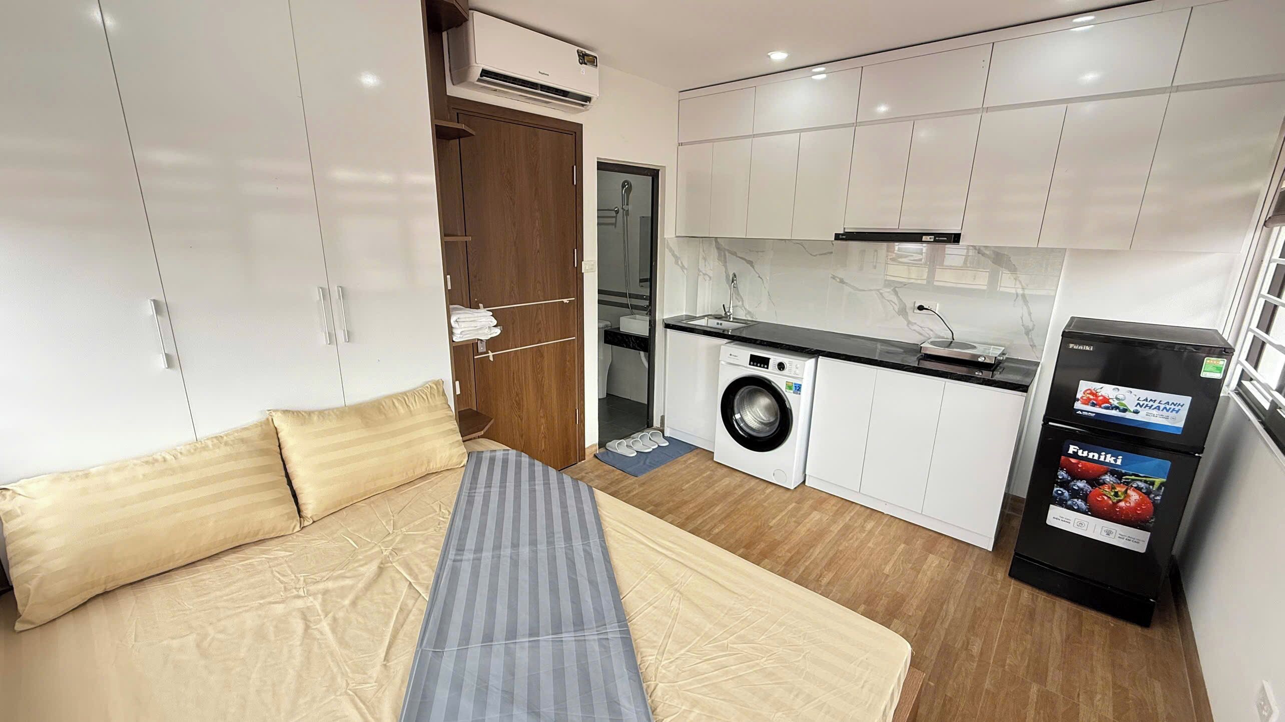A compact room with a bed, a washing machine, a kitchenette, and a small refrigerator.