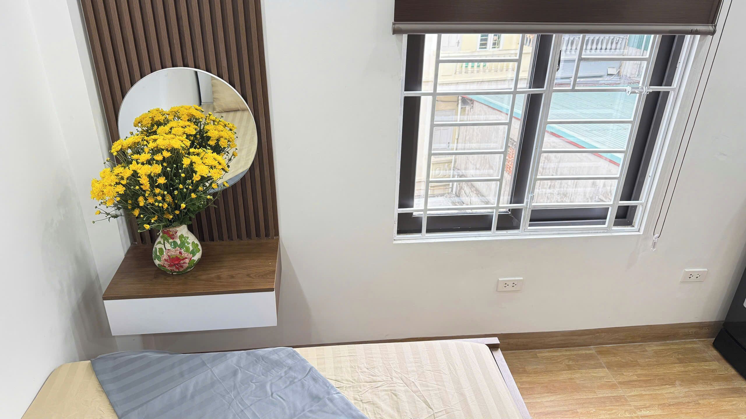 A bedroom with a bed, a window with a view of buildings, a mirror, and a vase with yellow flowers on a shelf.