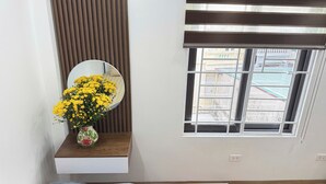 Un baño con espejo, ventana con vistas a edificios y repisa con un jarrón de flores amarillas.