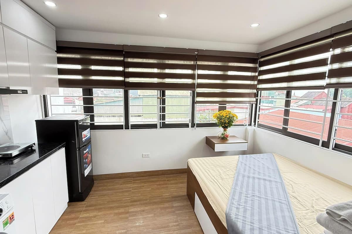 A compact bedroom with a bed, a small bedside table with a vase of flowers, a window with blinds, and a kitchenette with a refrigerator and a coffee machine.