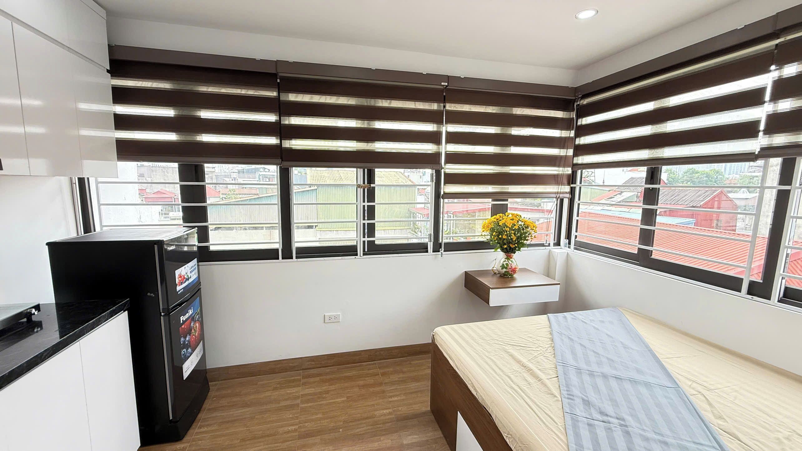 A small, modern room with a bed, a desk, and a refrigerator. There are large windows with blinds, and a view of buildings outside.