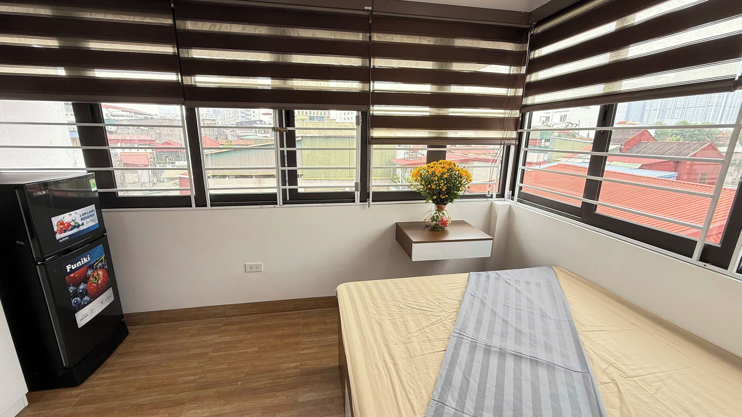 A hotel room with a bed, a small table with a vase of flowers, a minibar, and large windows with blinds.