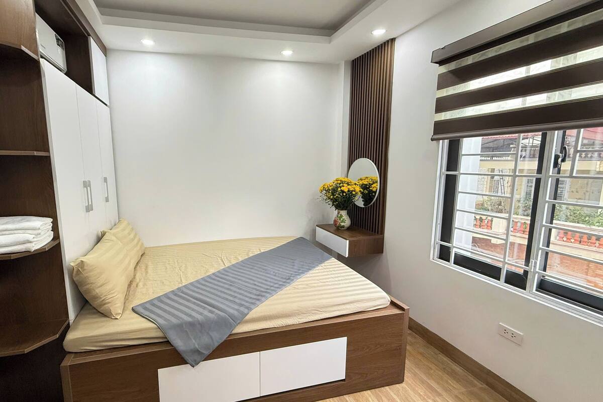 A bedroom with a bed, a wardrobe, a bedside table with a mirror and flowers, and a window with a view.
