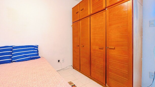 A bedroom with a bed, blue striped pillows, a wooden wardrobe, and a tiled floor. - Apto pet Friendly c Garagem em Rio Vermelho (Salvador)