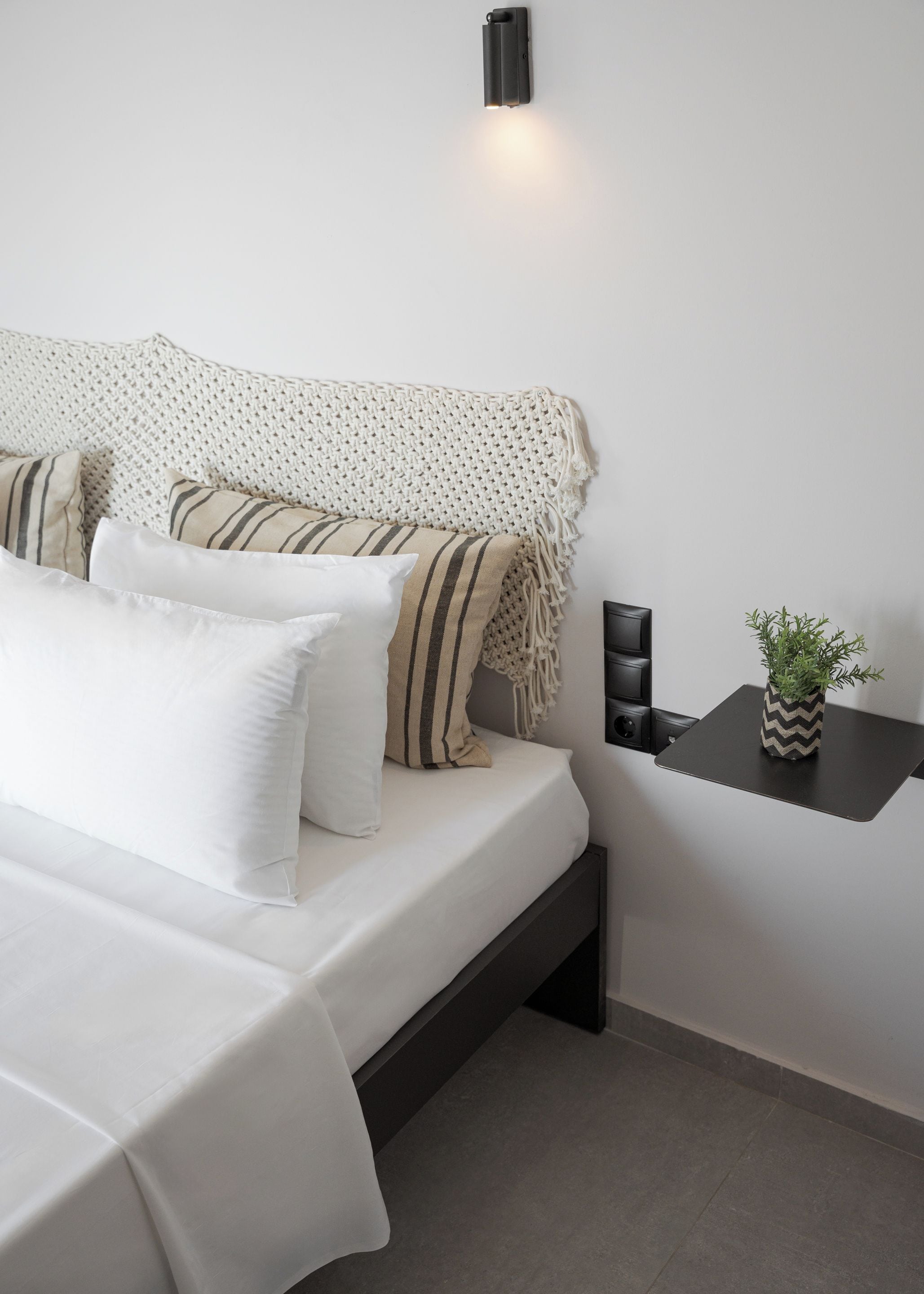 Une chambre moderne avec un lit, des oreillers, un plaid tricoté, une table de chevet avec une plante et une lampe fixée au mur.