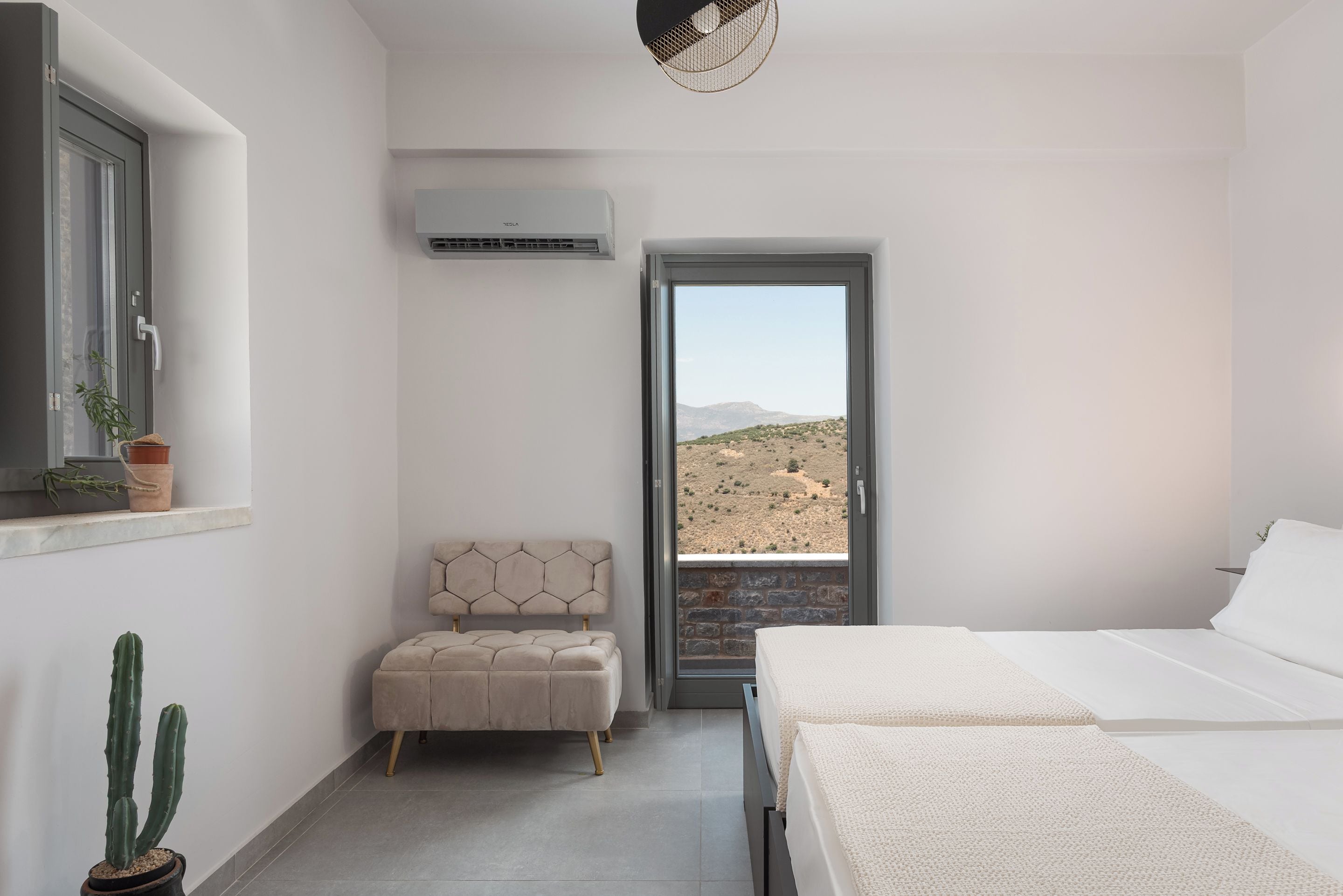 Un dormitorio con cama, ventana con vistas a un paisaje desértico, planta en maceta y unidad de aire acondicionado.