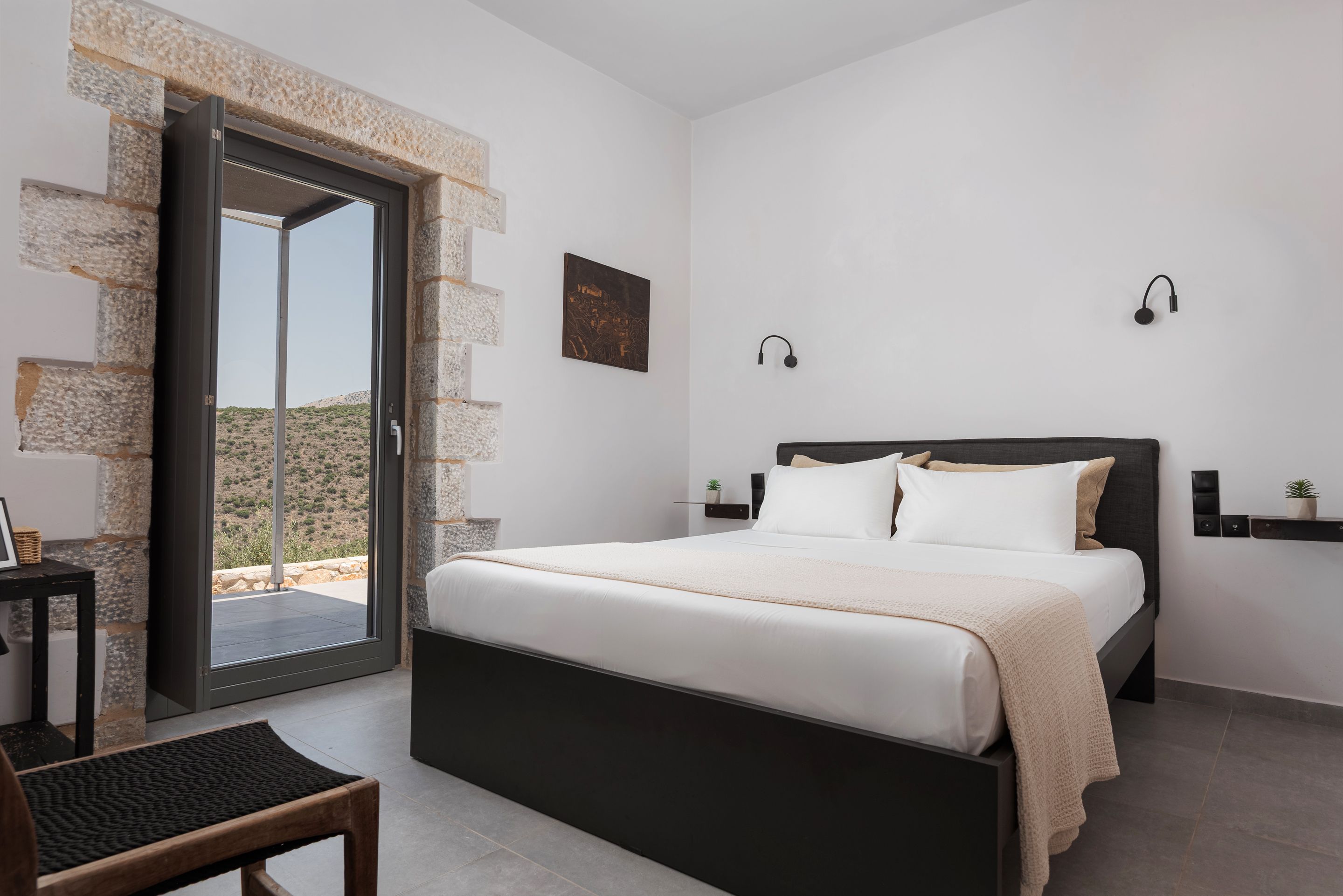 Quarto com cama, uma cadeira, mesinha, luminária na parede e vista através de uma porta de vidro.