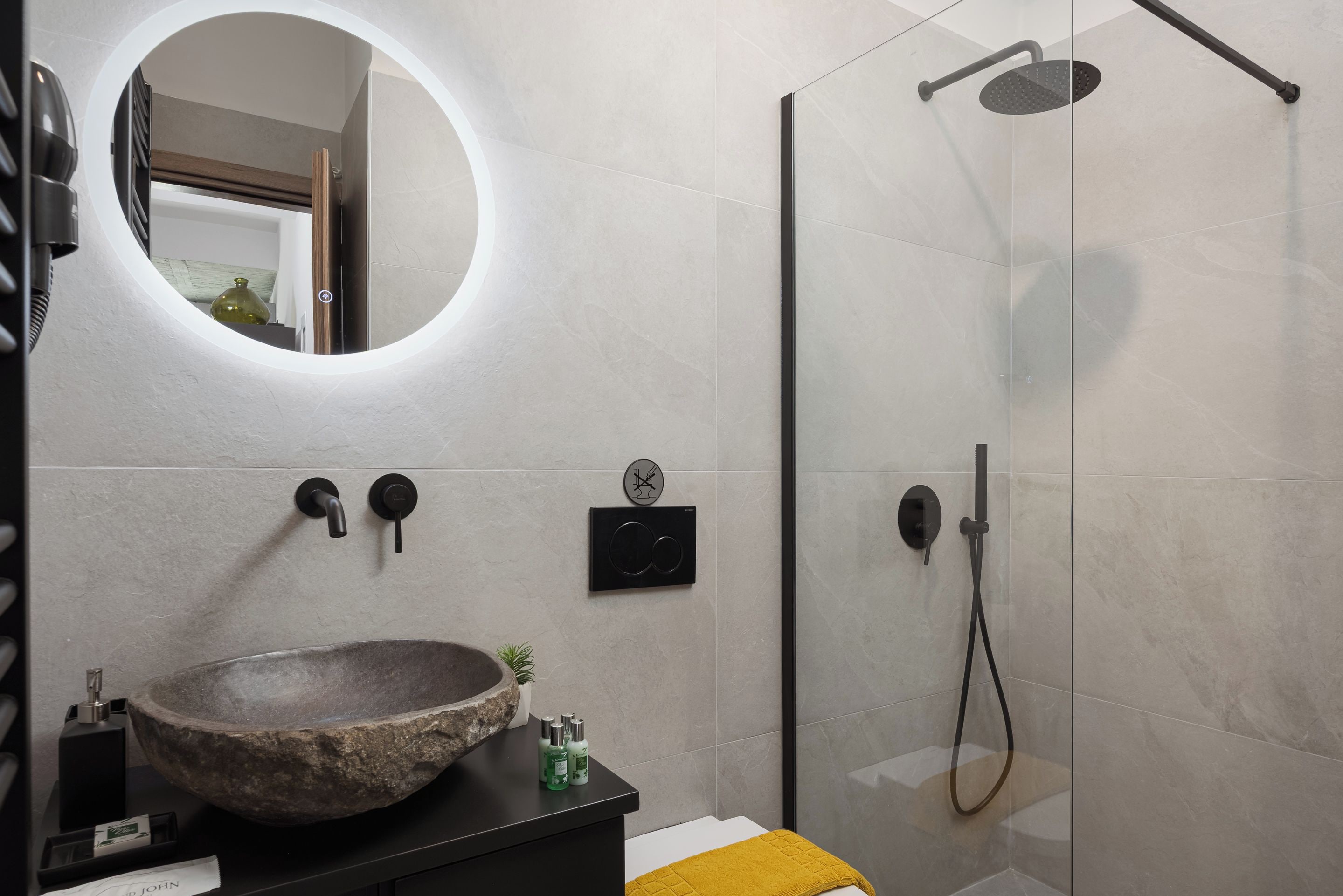 Een moderne badkamer met een ronde spiegel, een wastafel van natuursteen, een handdouche en een douche met glazen wand.