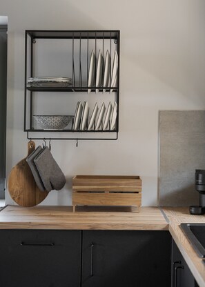 Une cuisine avec un plan de travail noir, une planche à découper en bois, un porte-ustensiles suspendu et une étagère en métal avec des assiettes et des bols.