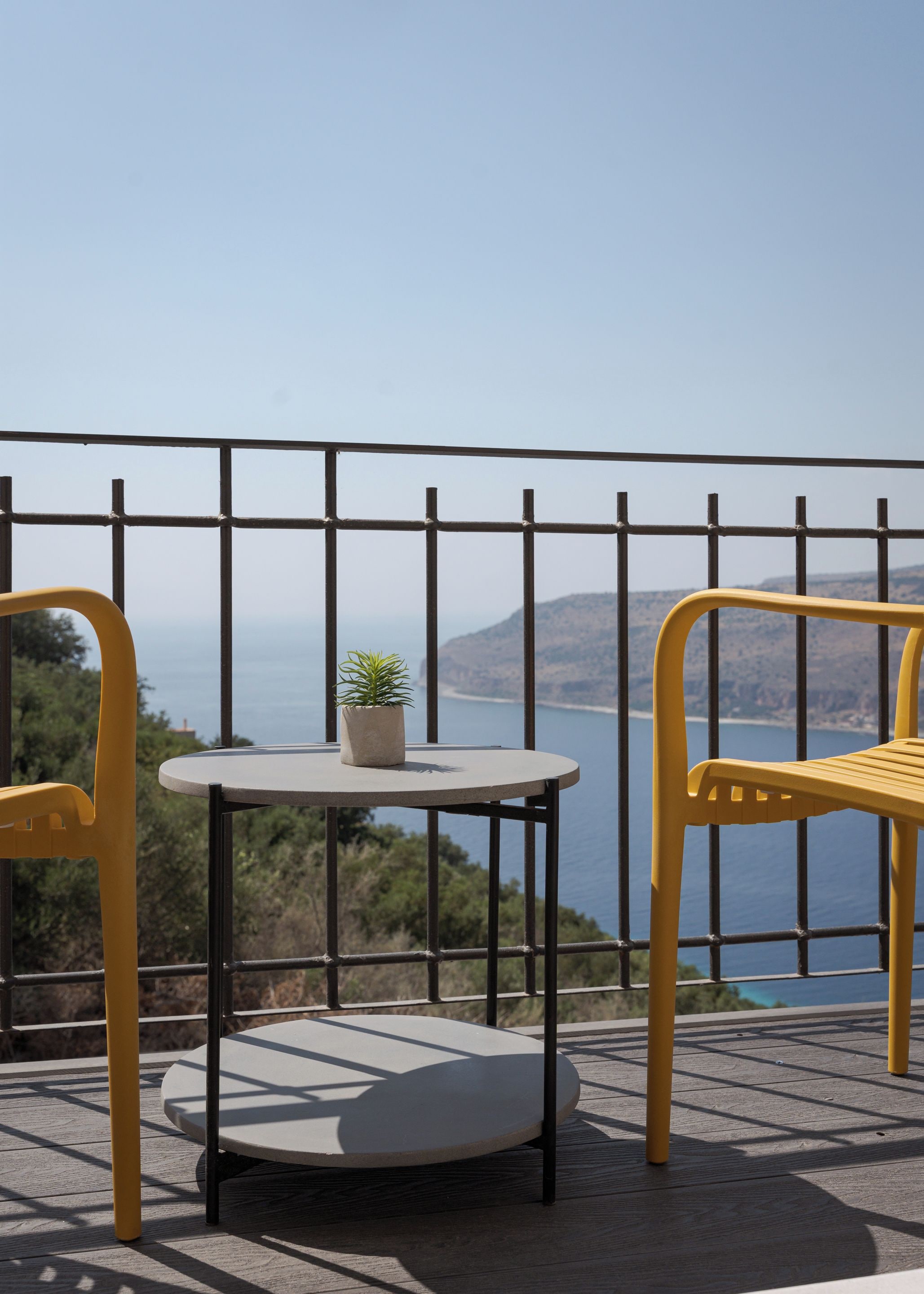 Un balcón con una mesa redonda y dos sillas amarillas, con vistas al agua y colinas.