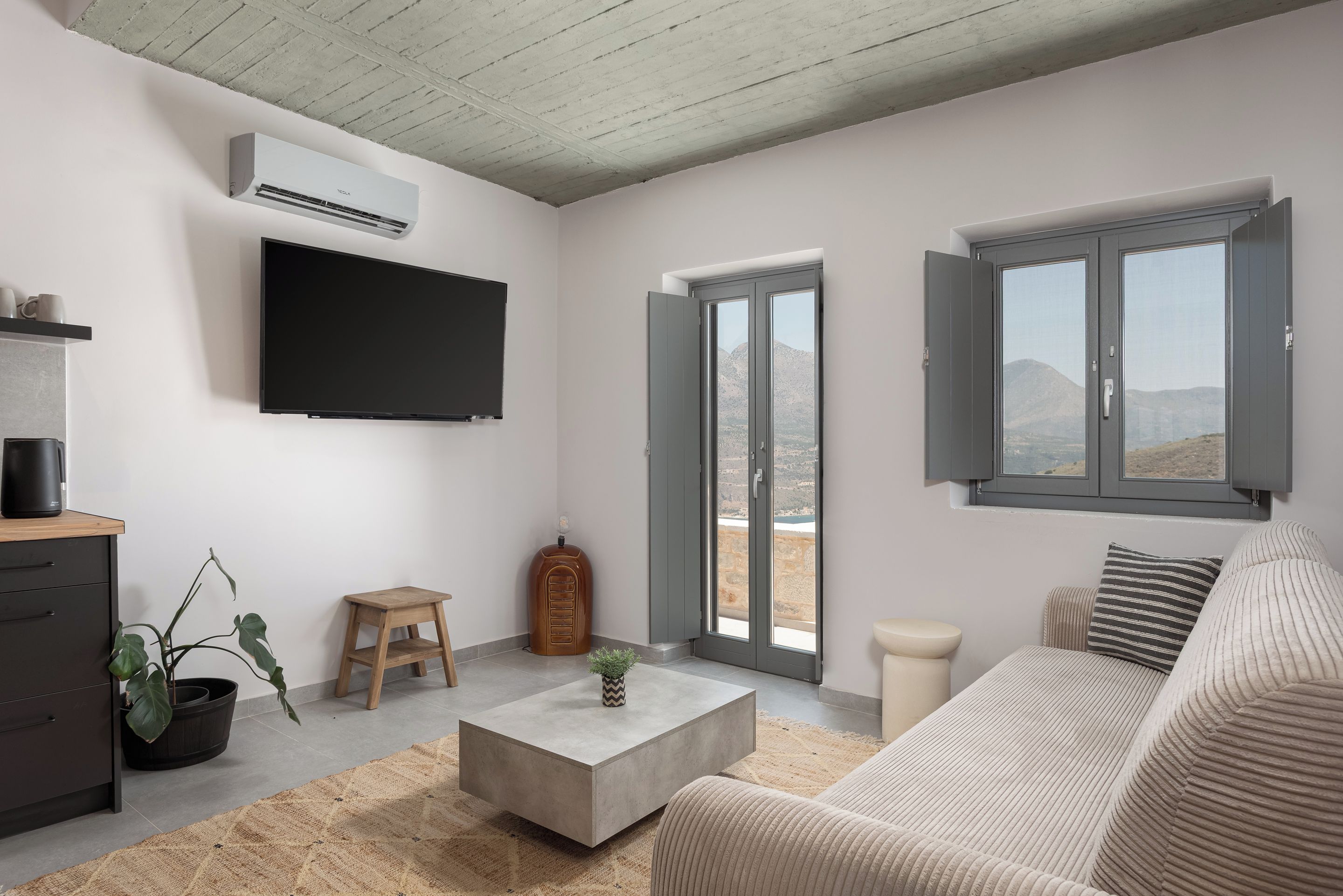 Ein modernes Wohnzimmer mit Flachbildfernseher, Blumentopf, Couchtisch, Sofa und einer Schiebetür zu einem Balkon mit Blick auf die Berge.