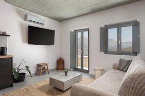 A modern living room with a flat-screen TV, a potted plant, a coffee table, a sofa, and a sliding glass door leading to a balcony with a mountain view.