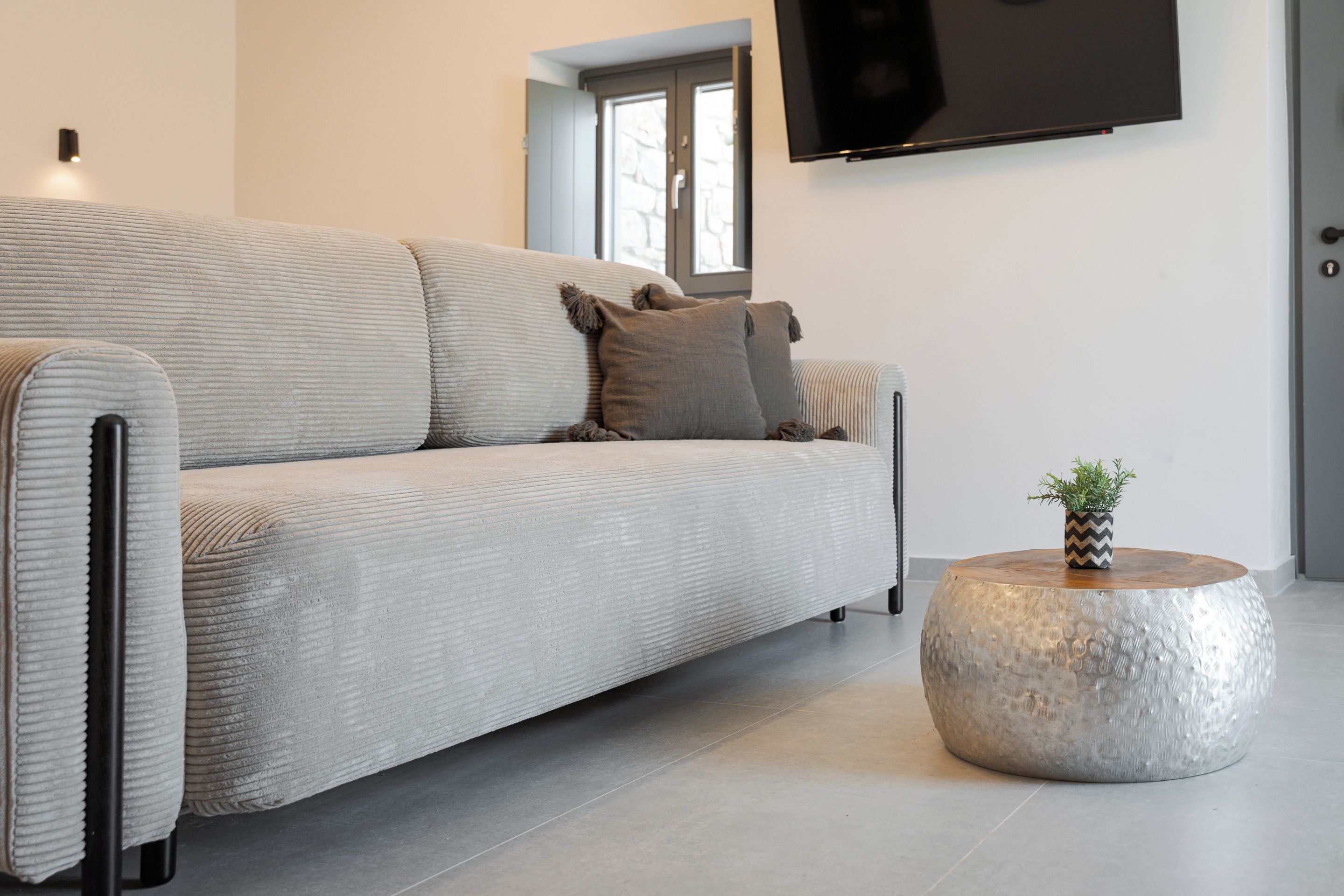 Een moderne woonkamer met een grijze bank, een ronde metalen salontafel en een flatscreen televisie aan de muur.