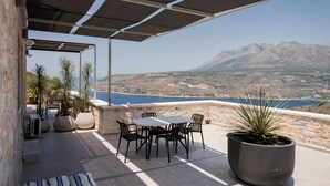 Una terraza con una mesa y sillas, plantas en macetas y vistas a un paisaje montañoso y al mar.